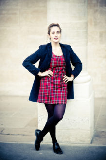 Clotilde, dressed in couleurs vives—a red plaid dress with black tights and shoes—stands confidently with hands on hips, leaning against a light stone column outdoors in the Jardin du Palais-Royal.