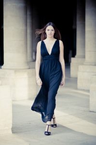 A woman with straight brown hair walks confidently outdoors at the Jardin du Palais-Royal, wearing a sleeveless, deep v-neck black dress and black heels. She is framed by stone columns and soft sunlight.