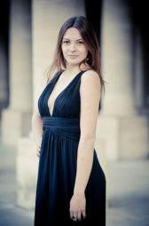 Anaïs, with long brown hair and a sleeveless black dress, stands outdoors at Palais-Royal, looking at the camera with a slight smile. The background features blurred stone columns and soft lighting.