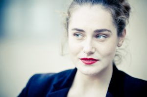 A woman with light skin, blue eyes, and red lipstick looks thoughtfully to the side. She has wavy brown hair pulled back, wearing a dark jacket. Softly blurred behind her are the couleurs of the Jardin du Palais-Royal.
