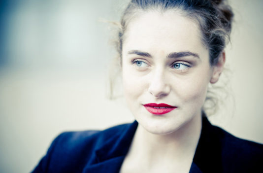 A woman with light skin, blue eyes, and red lipstick looks thoughtfully to the side. She has wavy brown hair pulled back, wearing a dark jacket. Softly blurred behind her are the couleurs of the Jardin du Palais-Royal.