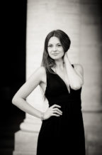 Anaïs poses in a sleeveless black dress before a grand column at Jardin du Palais-Royal, one hand on her hip and the other touching her neck, smiling subtly at the camera. The Noir et Blanc photo exudes timeless elegance.