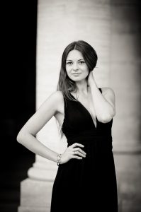 Anaïs poses in a sleeveless black dress before a grand column at Jardin du Palais-Royal, one hand on her hip and the other touching her neck, smiling subtly at the camera. The Noir et Blanc photo exudes timeless elegance.