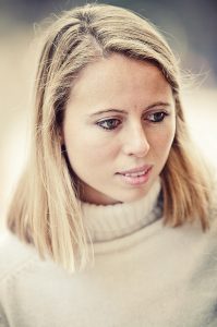 Barbara Welsh, a young woman with straight blonde hair and light eyes, looks downward in a white turtleneck sweater. The blurred background draws focus to her thoughtful expression.