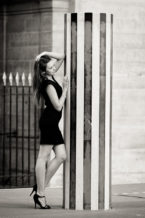 Anaïs, in a black dress and heels, strikes a chic pose with one hand on her head and the other on a striped pillar at Jardin du Palais-Royal. The photo captures the elegance of Noir et Blanc in an urban outdoor setting.