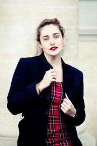 Clotilde, with wavy brown hair and red lipstick, stands confidently before a light stone column at the Jardin du Palais-Royal, wearing a dark blazer over a red plaid dress that highlights vibrant couleurs.
