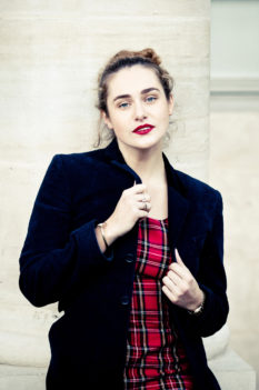 Clotilde, with wavy brown hair and red lipstick, stands confidently before a light stone column at the Jardin du Palais-Royal, wearing a dark blazer over a red plaid dress that highlights vibrant couleurs.