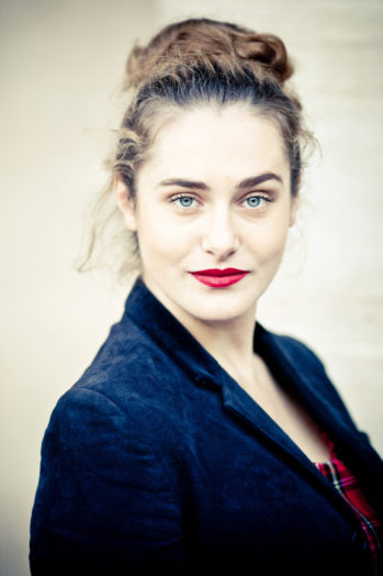 A woman with light skin, blue eyes, and red lipstick looks at the camera. She has brown hair tied up and is wearing a dark blazer over a red top, her Couleurs standing out against a light, neutral background reminiscent of Jardin du Palais-Royal.