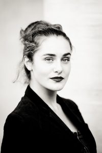Black-and-white portrait of Clotilde with light eyes and dark wavy hair pulled back, wearing a dark blazer. She looks confidently at the camera, set against a neutral background evoking the timeless elegance of Noir et Blanc.