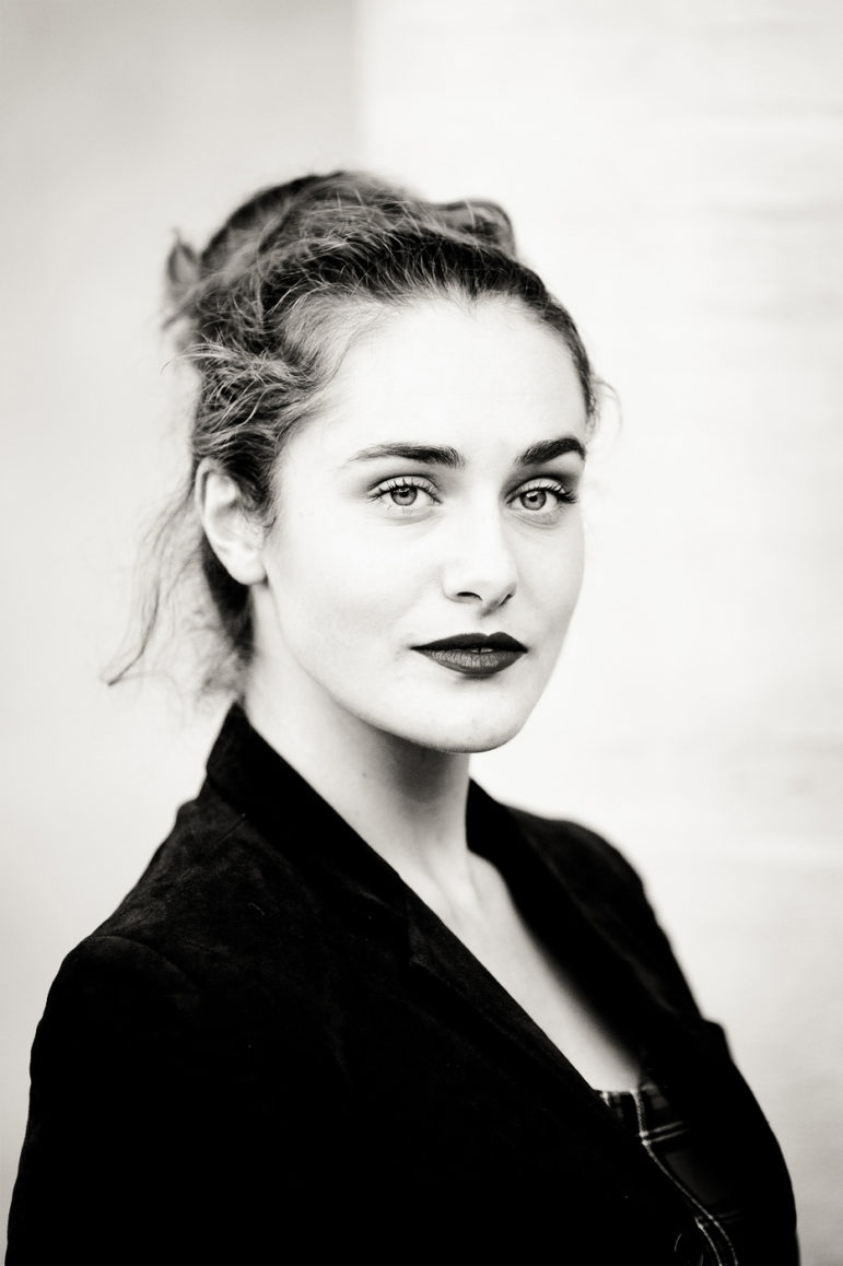 Black-and-white portrait of Clotilde with light eyes and dark wavy hair pulled back, wearing a dark blazer. She looks confidently at the camera, set against a neutral background evoking the timeless elegance of Noir et Blanc.