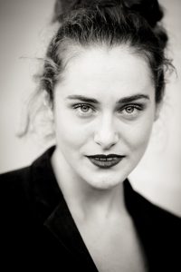 Noir et Blanc portrait of a woman with light eyes, curly hair pulled back, and dark lipstick. She is wearing a dark blazer and looking directly at the camera with a confident expression, echoing Clotilde at Jardin du Palais-Royal.