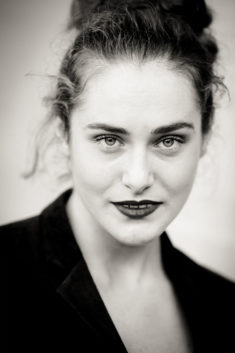Noir et Blanc portrait of a woman with light eyes, curly hair pulled back, and dark lipstick. She is wearing a dark blazer and looking directly at the camera with a confident expression, echoing Clotilde at Jardin du Palais-Royal.