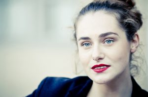 A woman with light skin, blue eyes, and red lipstick looks slightly to the side. Clotilde’s brown hair is tied back and she wears a dark blazer. The background, reminiscent of the Couleurs of Jardin du Palais-Royal, is blurred and light-colored.