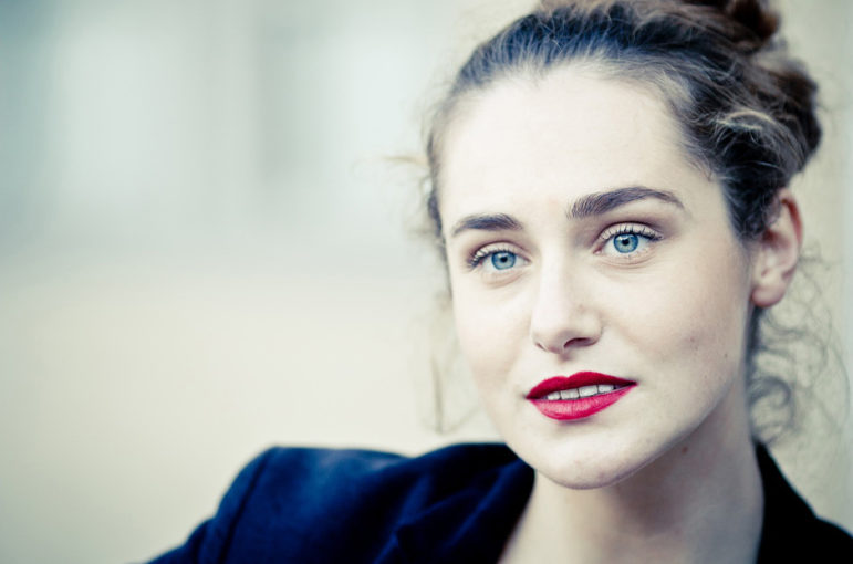 A woman with light skin, blue eyes, and red lipstick looks slightly to the side. Clotilde’s brown hair is tied back and she wears a dark blazer. The background, reminiscent of the Couleurs of Jardin du Palais-Royal, is blurred and light-colored.