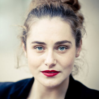 A woman with curly brown hair in a loose bun, wearing bright red lipstick and a black top, looks directly at the camera with a neutral expression and blue eyes. The softly blurred background hints at the Couleurs of Jardin du Palais-Royal.