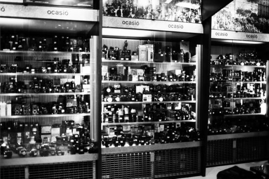 Black and white photo of glass display cases filled with cameras and equipment at Fotocasanova. Shelves are densely packed, with a sign above reading "ocasió," indicating this is a second-hand or used camera store.