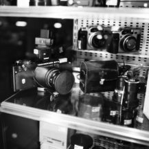 Black and white photo of a glass display case at Fotocasanova, featuring vintage and modern cameras, camera lenses, and a leather camera case arranged neatly on shelves. Reflections dance across the glass.