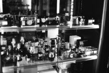 A black and white photo of a glass display case filled with various camera lenses, film canisters, and vintage cameras, arranged on multiple shelves in the Fotocasanova store or showroom.