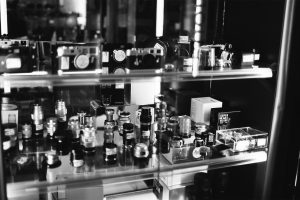 A black and white photo of a glass display case filled with various camera lenses, film canisters, and vintage cameras, arranged on multiple shelves in the Fotocasanova store or showroom.