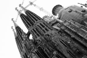 Black and white photo of the Sagrada Família in Barcelona, showing Gaudi’s tall, intricate towers and construction cranes against the sky. The angle is upward, highlighting architectural details.