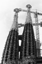 Two tall, ornate stone towers with intricate details rise beside a construction crane against a cloudy sky; the towers are part of Gaudi's unfinished masterpiece, the Sagrada Familia in Barcelona.