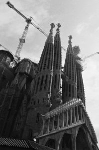 Black and white photo of Gaudi’s Sagrada Familia in Barcelona, showing its tall, ornate spires and cranes above, highlighting ongoing construction against a partly cloudy sky.