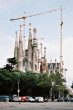 The Sagrada Familia basilica in Barcelona, designed by Antoni Gaudi, is seen under construction with cranes towering above its spires. Trees, cars, and pedestrians fill the lively street in the foreground.