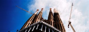 A panoramic view looking up at the Sagrada Familia in Barcelona, showcasing Gaudi’s tall, ornate towers against a blue sky with clouds. Construction cranes are visible beside the unfinished basilica.