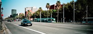 A city street with light traffic, including a black car in the foreground and a green tram in the background, lined with palm trees and modern buildings under a clear blue sky.