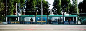 A modern green and white tram stopped at a station with several people waiting on the platform, surrounded by tall trees and clear skies in the background.