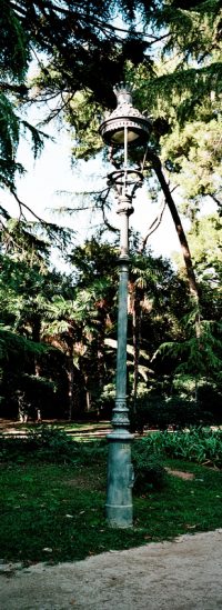 A tall, ornate street lamp stands on a grassy area by a dirt path in Parc de Pedralbes, Barcelona, surrounded by lush green trees and foliage in a sunlit park.