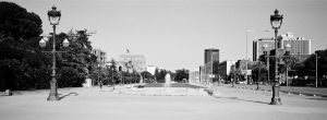 Wide boulevard with old-fashioned street lamps, empty pavement, a central fountain, and modern buildings in the background under a clear sky. Trees line the street on the left, while palm trees and more buildings are on the right.