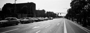 A wide city street with multiple lanes and white arrows, lined with cars waiting at a red light on the left. The right lane is mostly empty, with trees and buildings along the sidewalks.