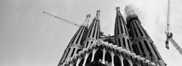 Black and white photo of Gaudi’s iconic Sagrada Familia in Barcelona, showcasing its ornate spires with cranes in the background, highlighting ongoing construction against a mostly clear sky.