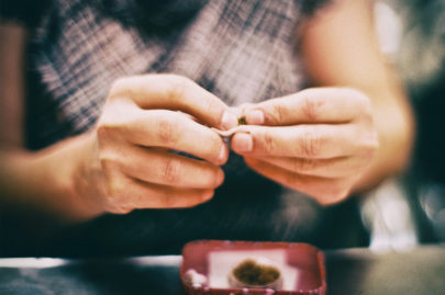 A person’s hands are rolling something small, possibly a cigarette or joint, over a table with a red tray containing loose material. The scene, reminiscent of quiet errance, is focused on the hands and tray while Cambrils blurs in the background.