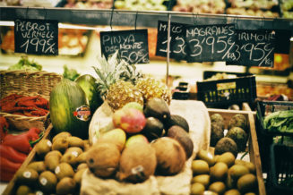 A vibrant market display in Cambrils showcases assorted fruits—pineapples, avocados, coconuts, passion fruits, and red peppers—with hand-written price signs in Spanish capturing the spirit of errance and local life.