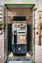 A close-up view of a weathered public payphone in a glass booth on a city street in Cambrils, with a numeric keypad, coin slot, and handset visible. Buildings and a streetlamp blur into the background, evoking an air of quiet errance.