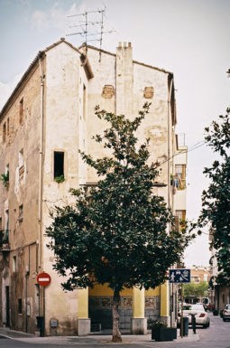 A tall, weathered building stands on a street corner in Cambrils, with a leafy tree in front. The faded facade hints at quiet errance, antennas on the roof, shuttered windows, and a red no-entry traffic sign on the left.