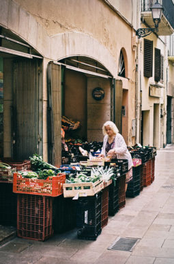 An elderly woman arranges fresh fruits and vegetables in crates outside a small market on a narrow, sunlit street in Cambrils, lined with old buildings.