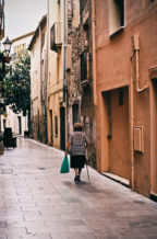 An elderly woman with a cane and a green shopping bag walks alone down a narrow, empty street in Cambrils, her quiet errance framed by old buildings in warm tones.