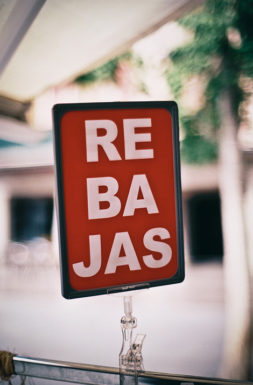 A red sign with bold white letters spelling "REBAJAS"—hinting at irresistible discounts—stands outdoors in Cambrils, with a blurred background of trees and buildings lending a touch of urbane errance to the scene.