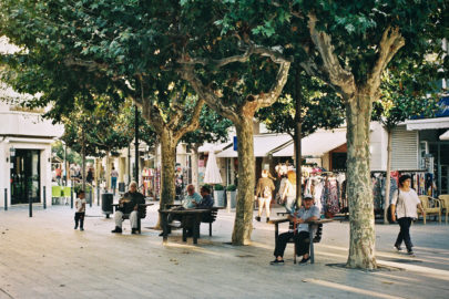 A tree-lined street in Cambrils with people sitting on benches and walking. Shops with clothes and goods are visible in the background, sunlight filtering through the leaves, casting dappled shadows that invite a gentle errance along the pavement.