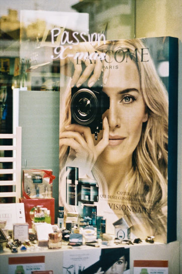 A store display in Cambrils showcases a large Lancôme advertisement featuring a woman holding a camera, surrounded by various beauty products and perfumes arranged on shelves below the poster, evoking a sense of errance and discovery.