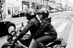 Two people wearing helmets and jackets ride a motorcycle through a city street, embodying the spirit of Génération motard. The passenger looks toward the camera while shops and buildings line the black and white background.
