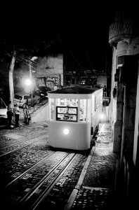 A vintage tramway illuminated at nuit travels up a steep, cobblestone street in Lisbonne, lined with buildings and graffiti walls; a few people walk nearby under streetlights.