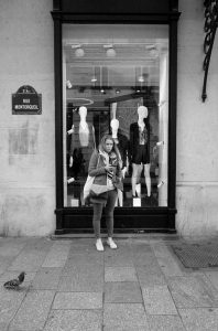 A young woman stands on the sidewalk in front of Jeanne Papillon's fashion store window, displaying mannequins dressed in various outfits. She is looking down at her phone, wearing a jacket and scarf. A pigeon is on the pavement near her. A "Rue Montorgueil" street sign is visible.