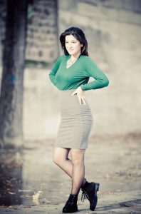 Anne-Sophie Sénéquier, with long dark hair, stands outdoors, smiling slightly. She wears a green long-sleeve top tucked into a gray knee-length skirt and black boots. The background of blurred trees and a stone wall adds to the autumnal feel with leaves scattered on the ground.