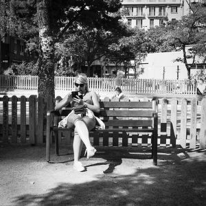 Miranda Perez sits on a park bench, reading her phone as sunlight casts gentle shadows. She wears sunglasses and casual clothes. Behind her, a fenced playground is framed by trees and distant buildings.