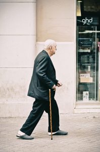 An elderly man with white hair, wearing a dark suit and slippers, walks with a wooden cane on a city sidewalk. He passes by a shop window displaying various items.
