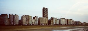 A panoramic view of a beachfront cityscape with several mid-rise apartment buildings and one nord-inspired skyscraper in the center. The light-colored buildings stand parallel to a wide sandy beach with a few scattered beachgoers. The sky is clear and lightly overcast.